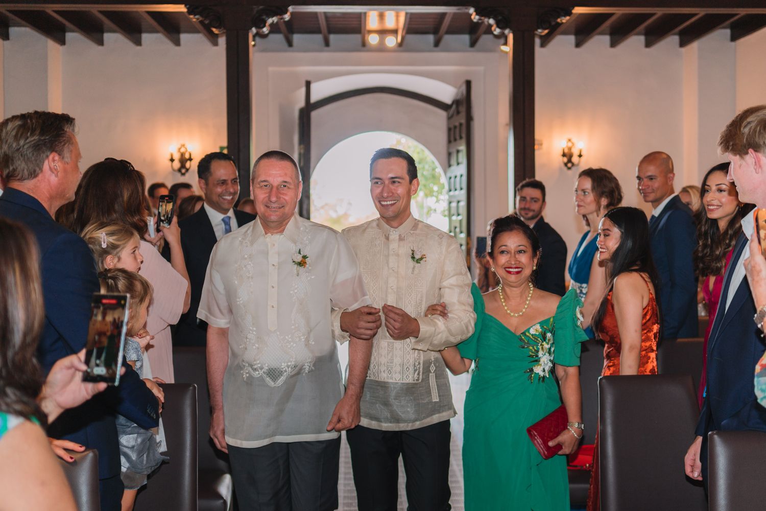 A group of wedding guests and participants gather inside a venue with elegant decor and archways.