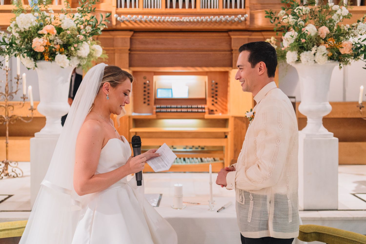Bride and groom exchanging vows in front of an ornate church organ decorated with elegant white floral arrangements.