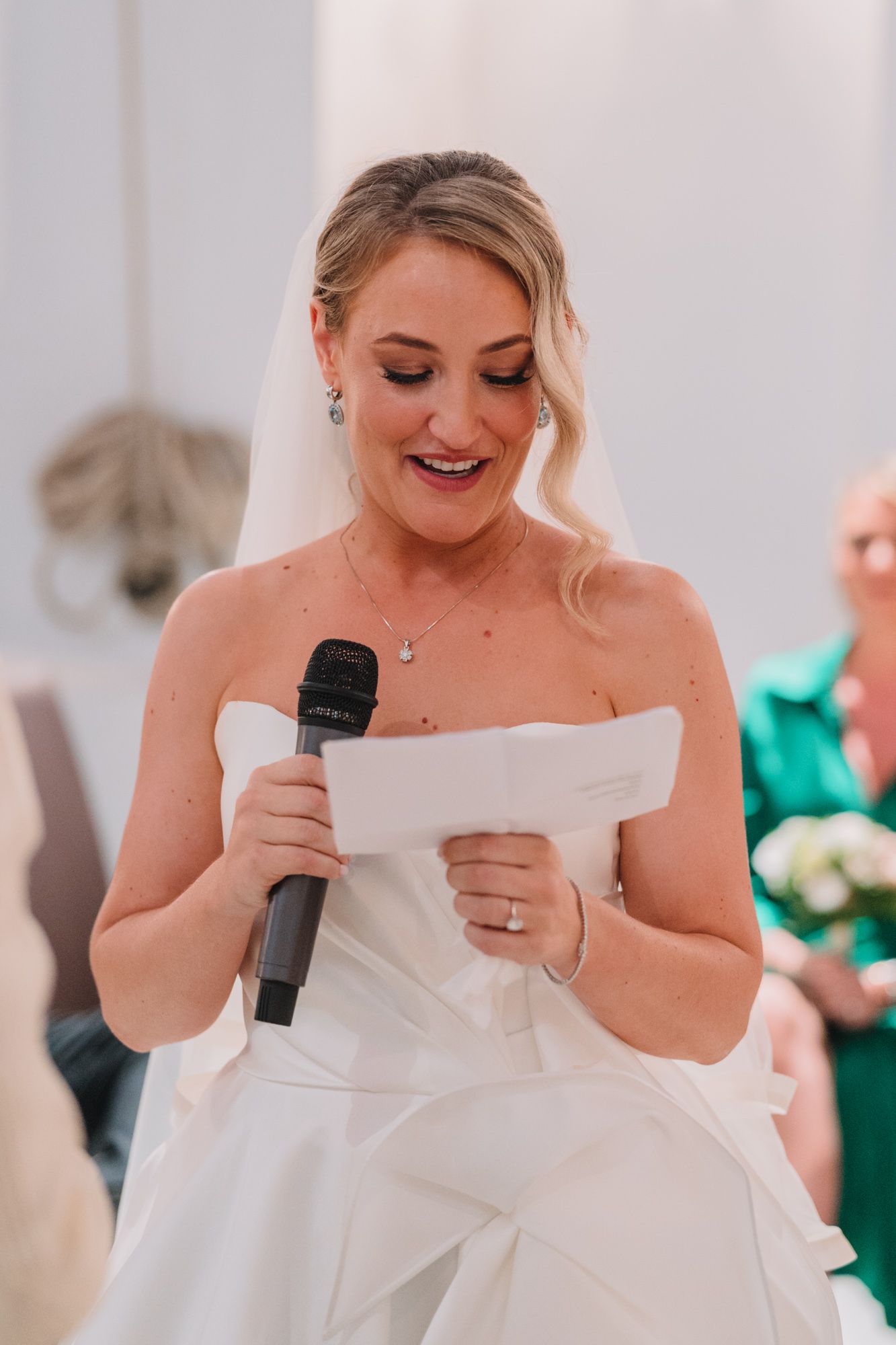 A bride in a white wedding dress reads from paper while holding a microphone during the reception.