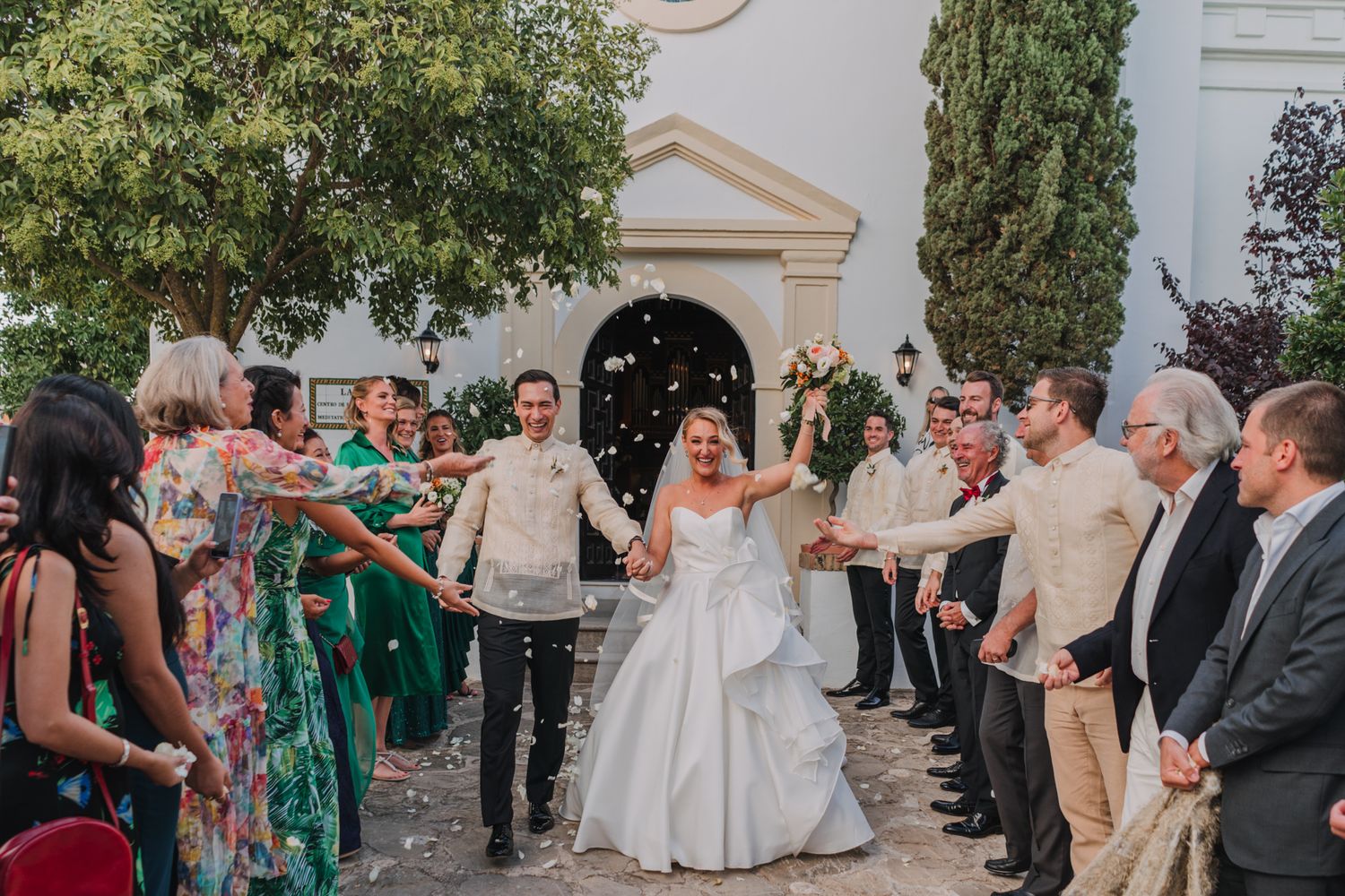 Wedding guests form an aisle to celebrate the newlyweds outside a white building with confetti.