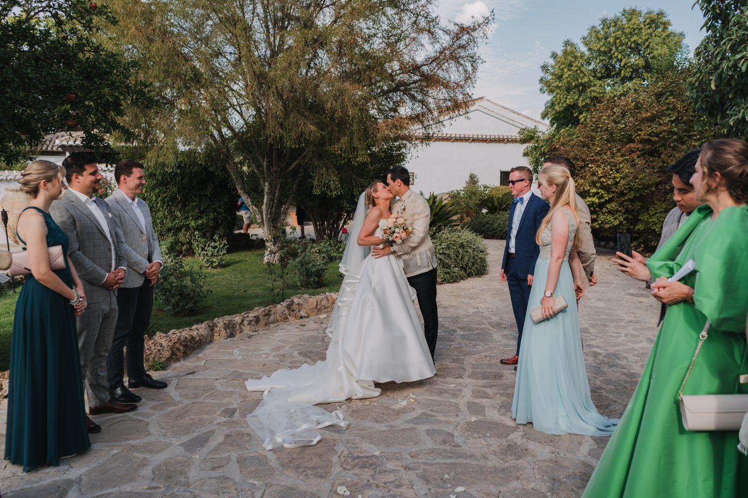 Wedding party and guests gather on a stone pathway during an outdoor ceremony.