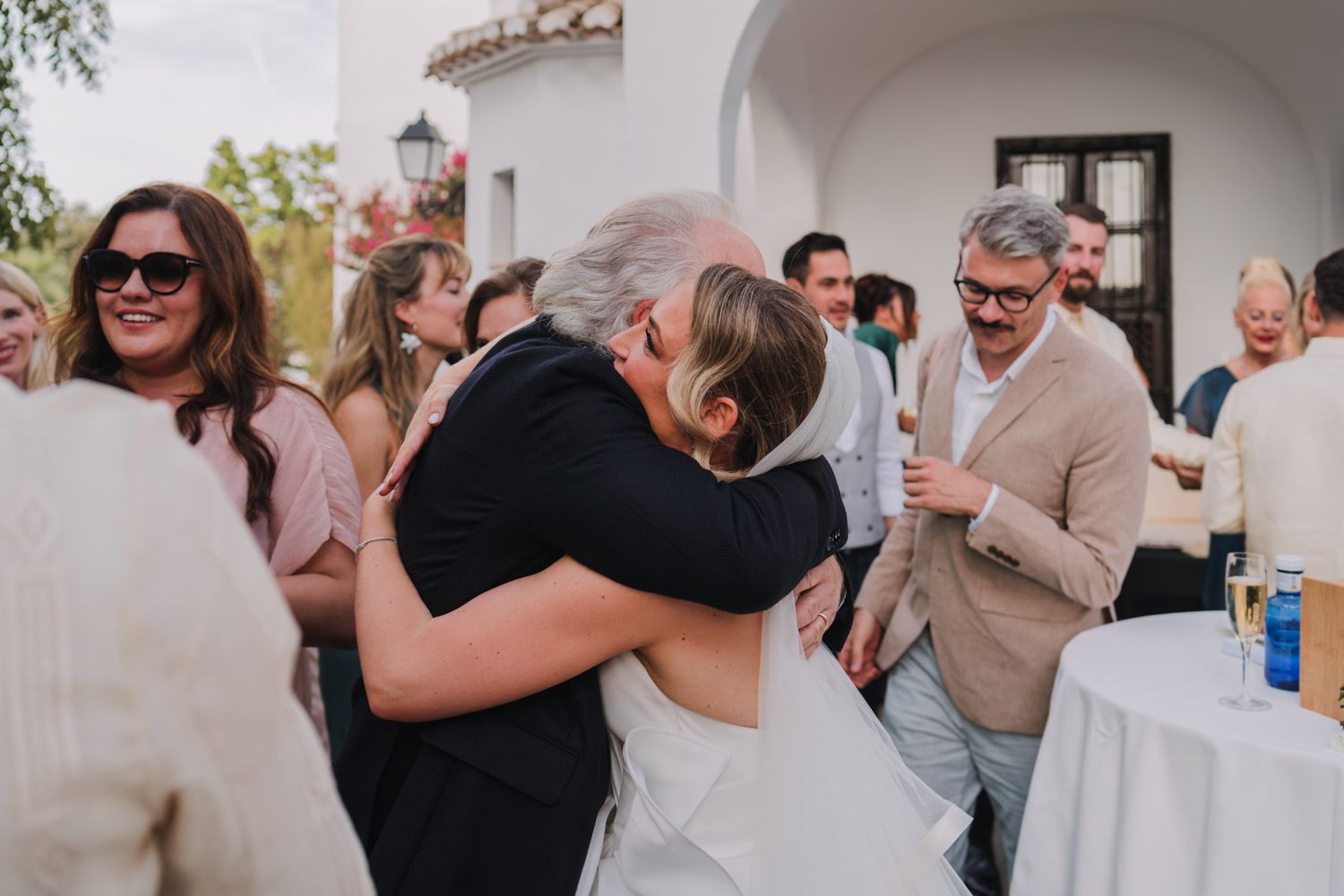 Wedding guests gather during an outdoor reception celebration with hugs and embraces under arched architecture.