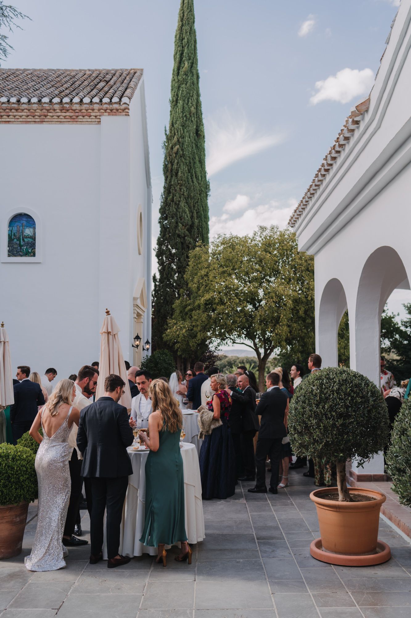 Wedding guests gather in an elegant Spanish-style courtyard with white stucco walls and cypress trees.