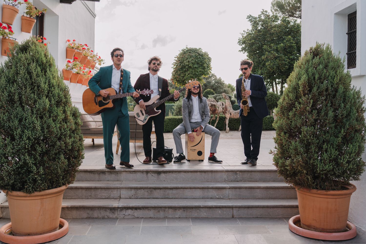 Five-piece band with guitars and instruments poses on stone steps between potted topiary plants.