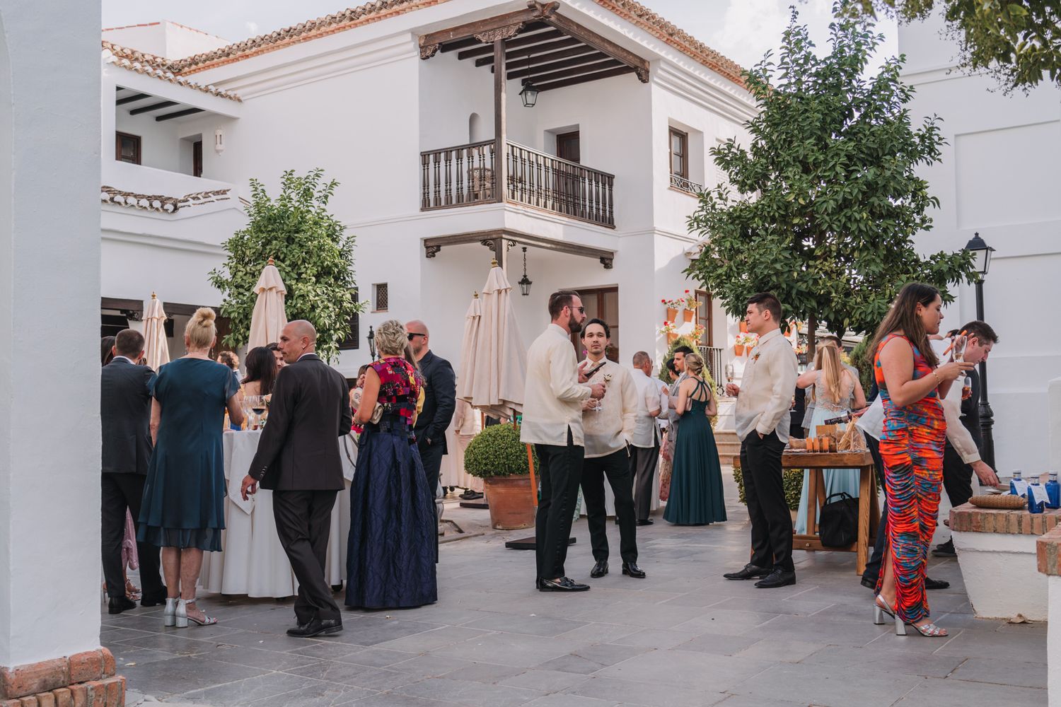 Wedding guests socializing in a Mediterranean-style courtyard with white buildings.