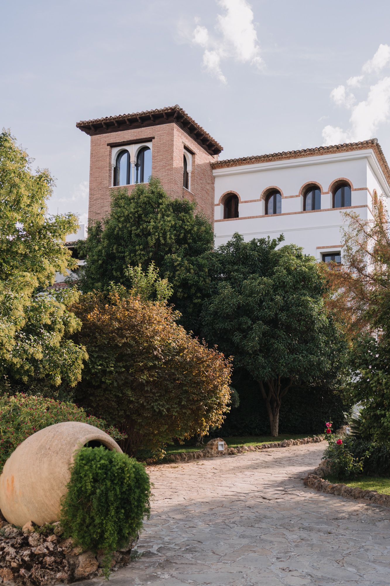 Spanish colonial style building with tower and arched windows surrounded by lush greenery and curved pathway.