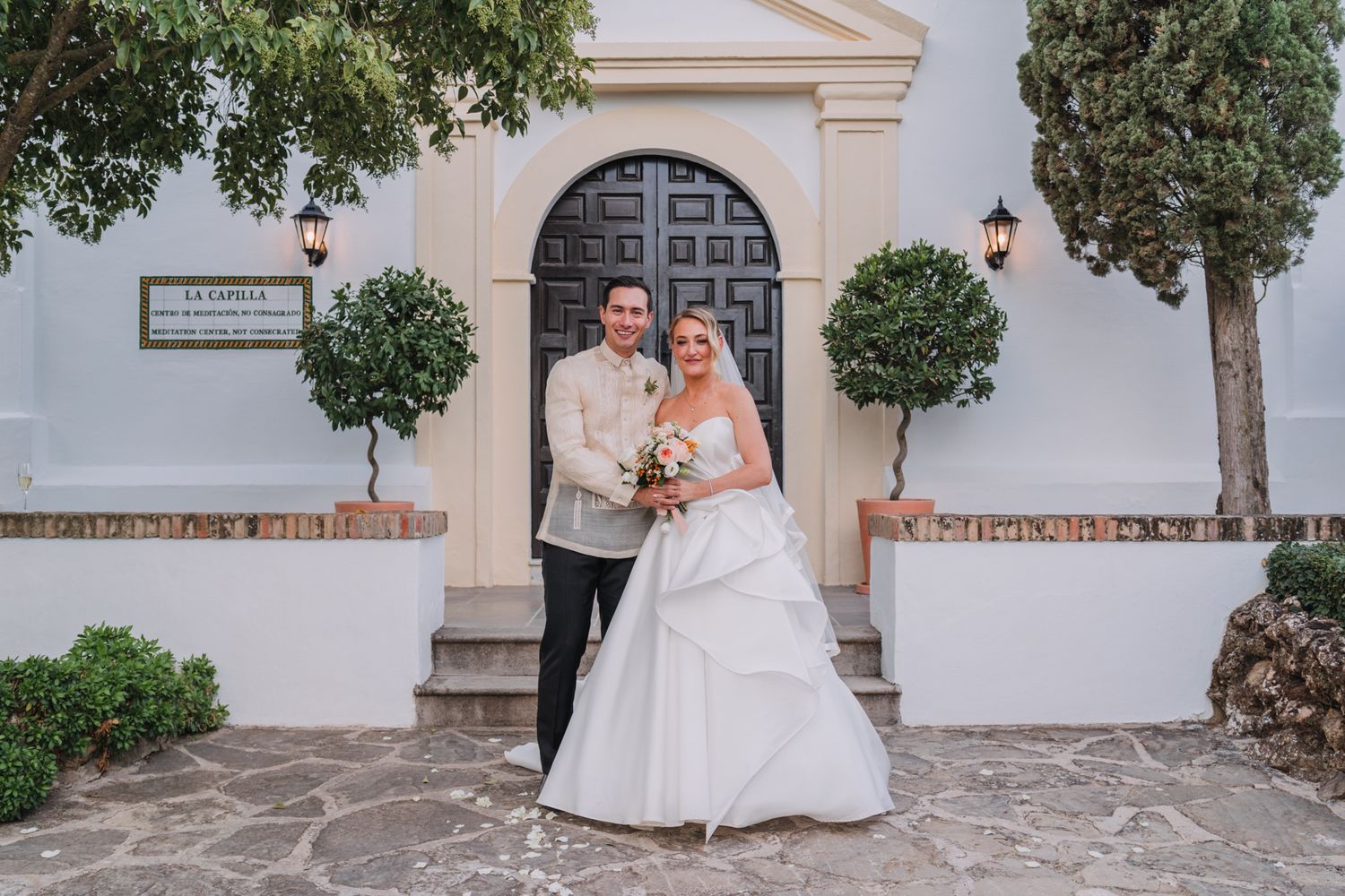 Elegant wedding portrait in front of a white Mediterranean church entrance with decorative topiary.