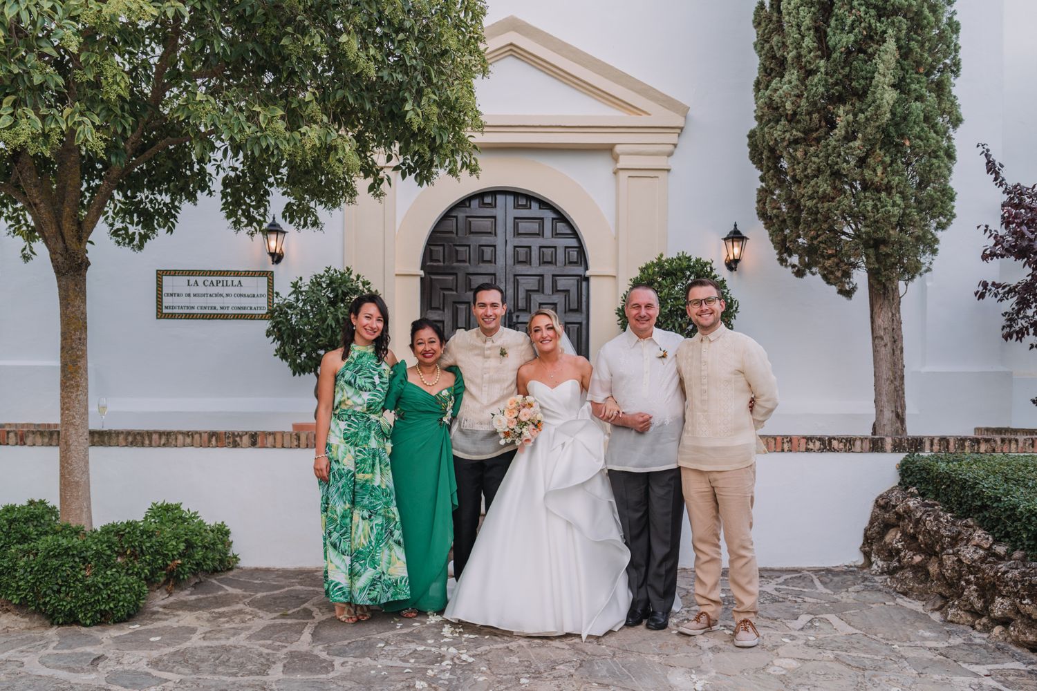 Wedding party in green and cream attire poses together outside a traditional white church building.