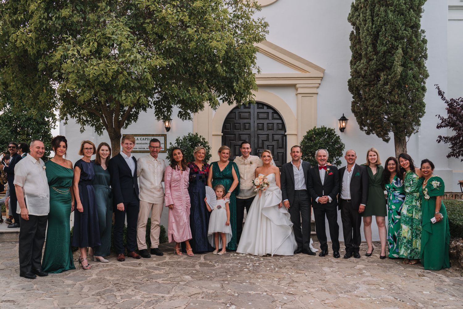 Large wedding party group photo gathered outside a Mediterranean church entrance with cypress trees.