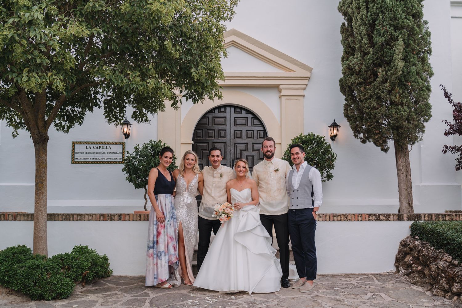 Small wedding party poses in front of white church wall with ornate doorway and cypress trees.