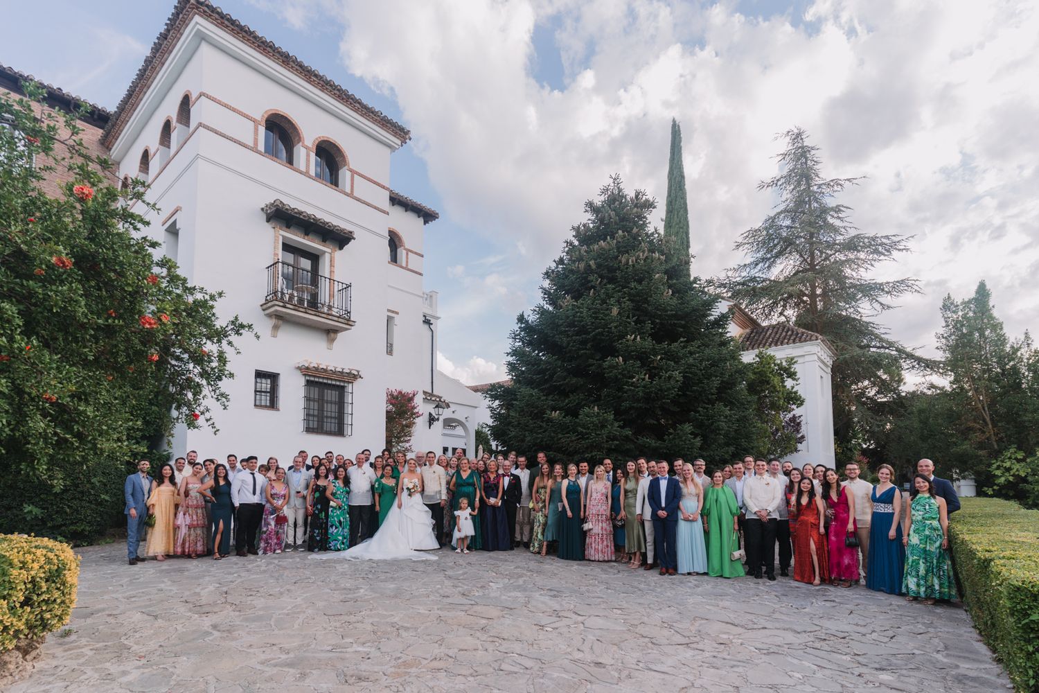 Large wedding party group photo in front of a white Spanish-style villa with tower and cypress trees.