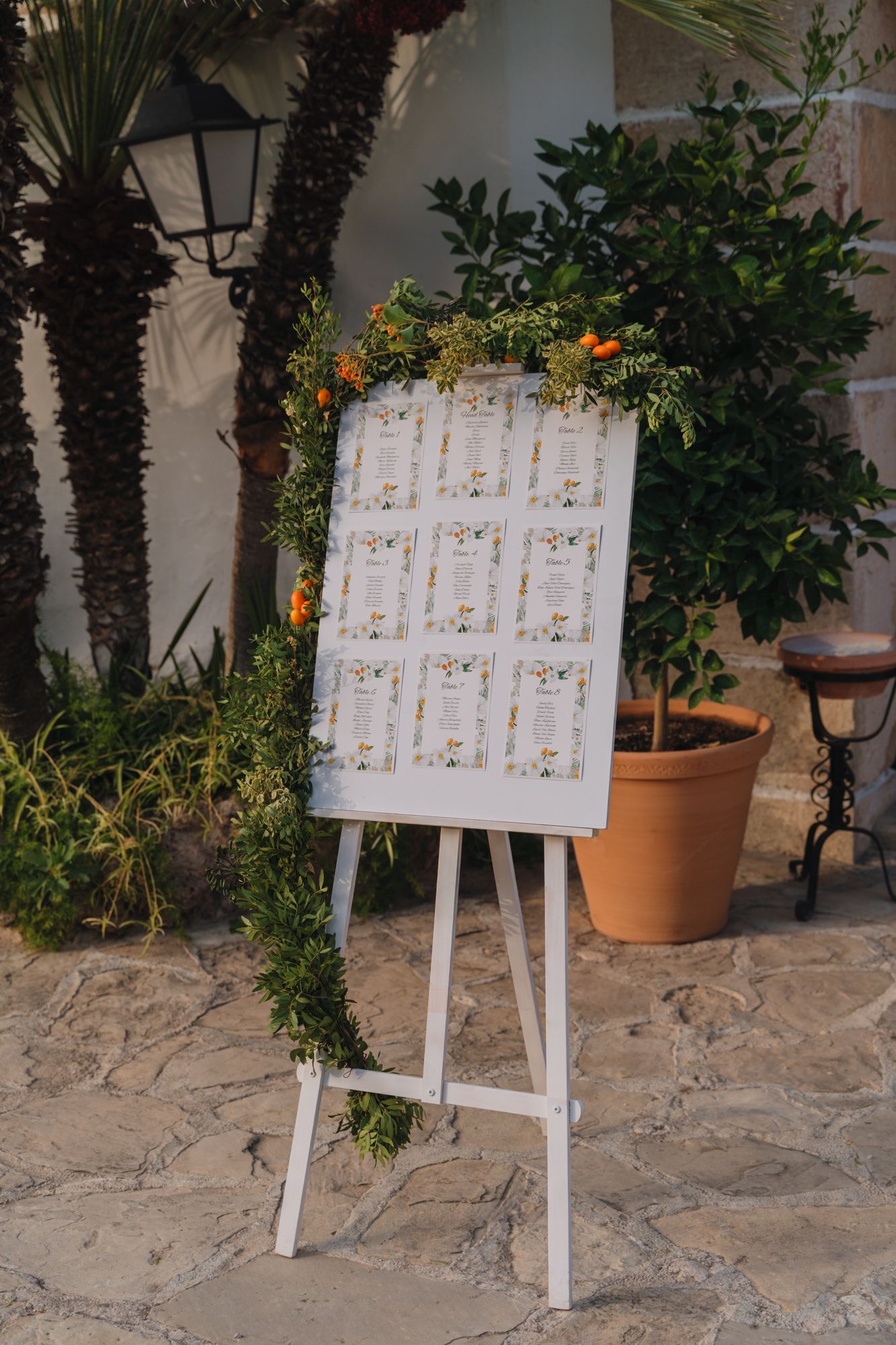 A white wedding seating chart display on an easel decorated with greenery and flowers against a stone patio wall.