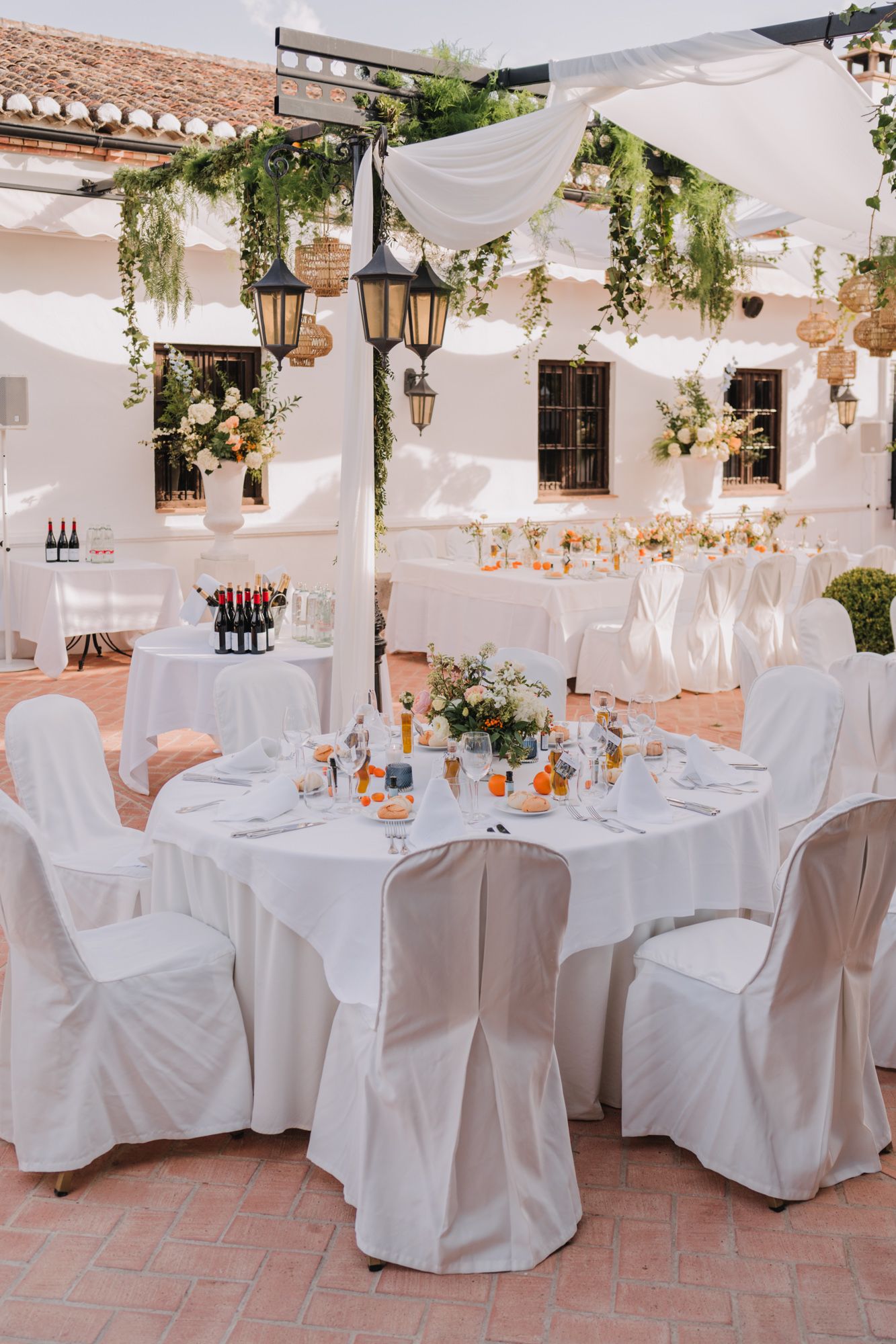 Romantic outdoor wedding reception setup with white draped tables and hanging greenery.