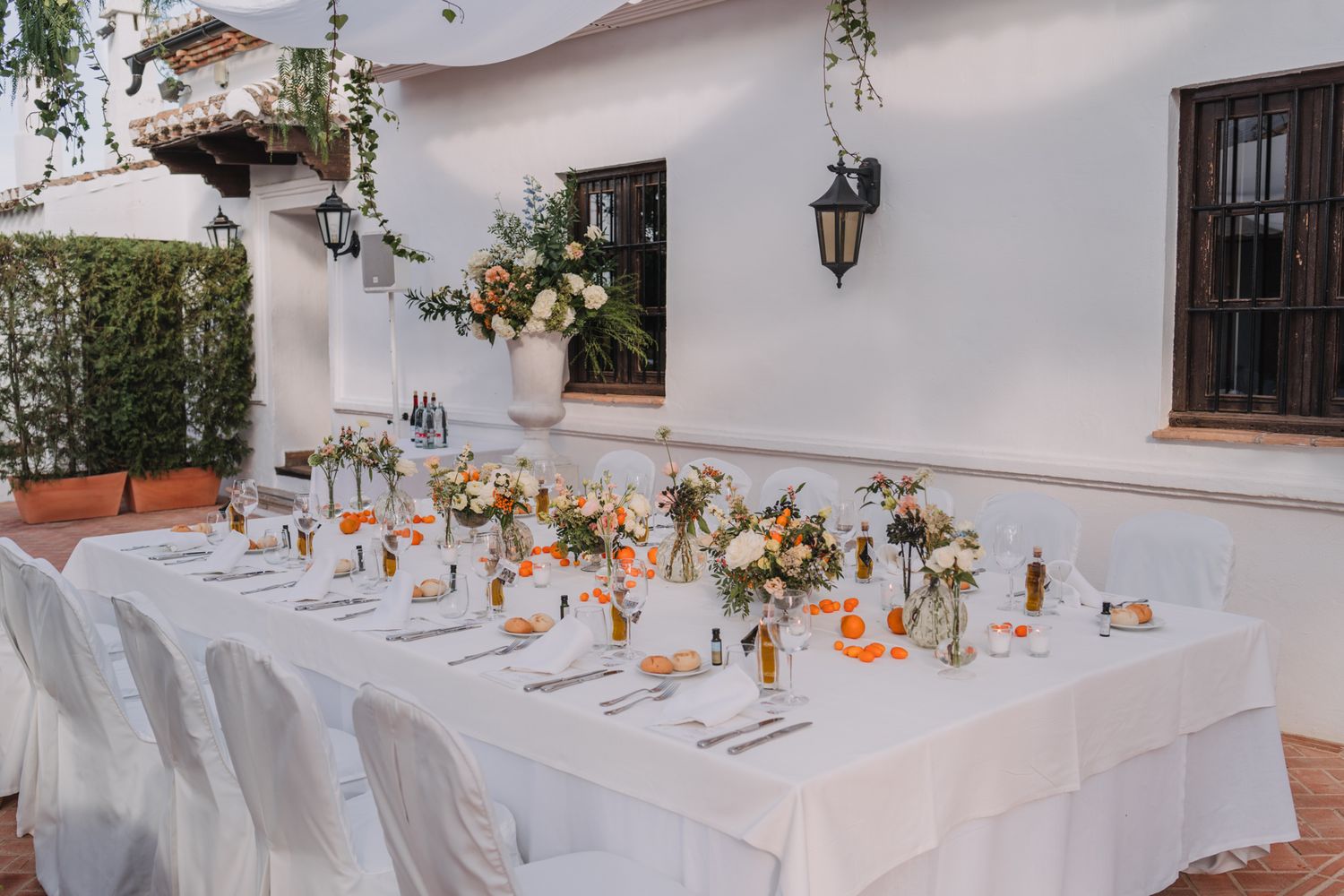 Long white reception table set with floral arrangements and place settings against stucco wall.