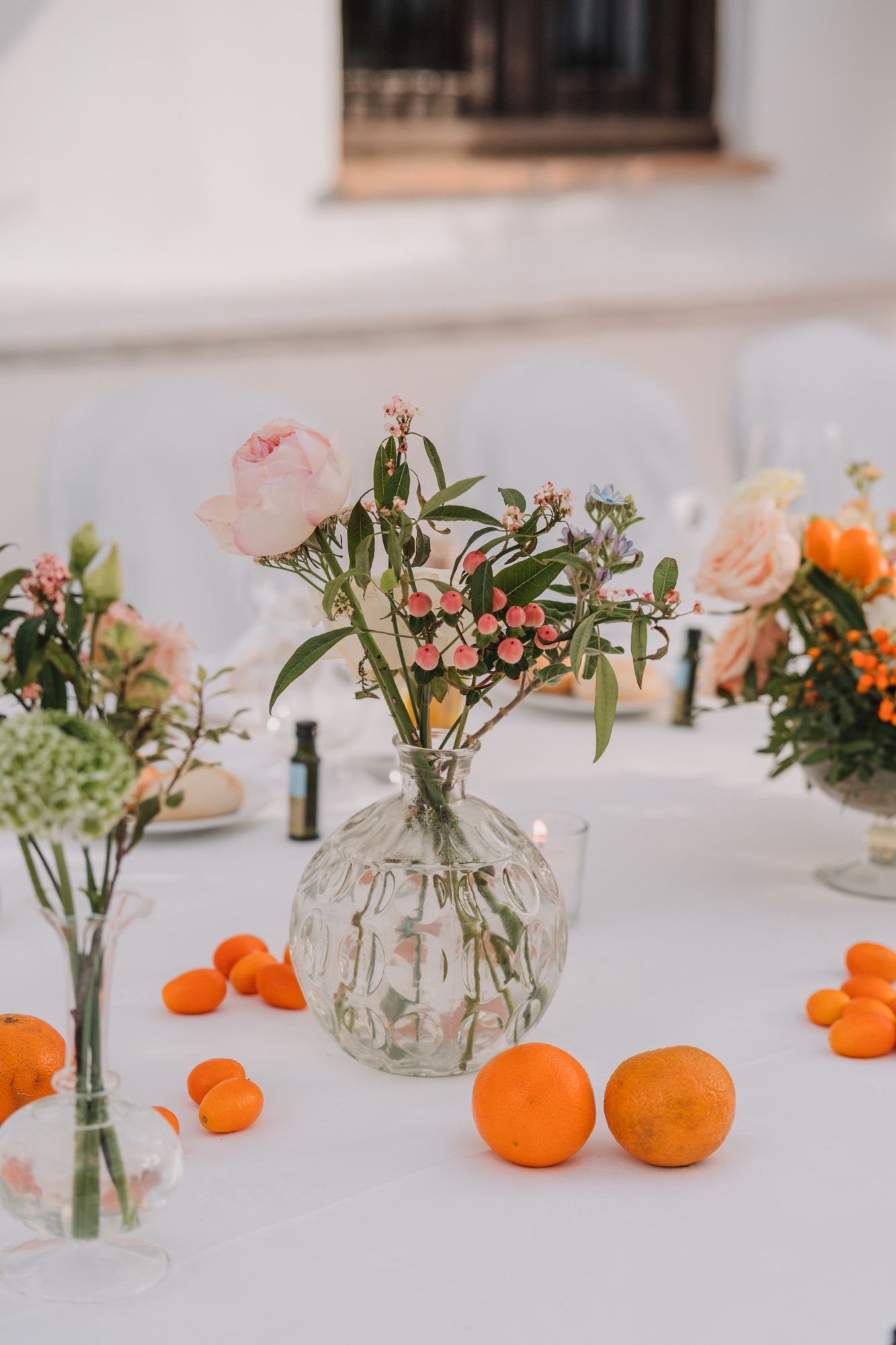 Wedding table centerpieces featuring pink roses in glass vases with fresh oranges scattered on a white tablecloth.