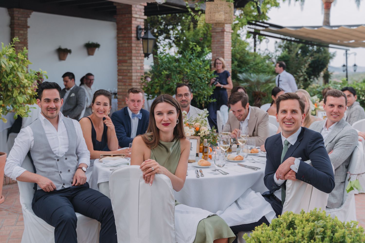Wedding guests sit at a round table with white linens during an outdoor reception at a Mediterranean-style venue.