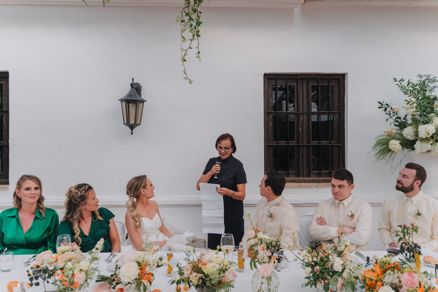 Wedding party gathered at an elegant outdoor reception table with speeches and decorative floral centerpieces.