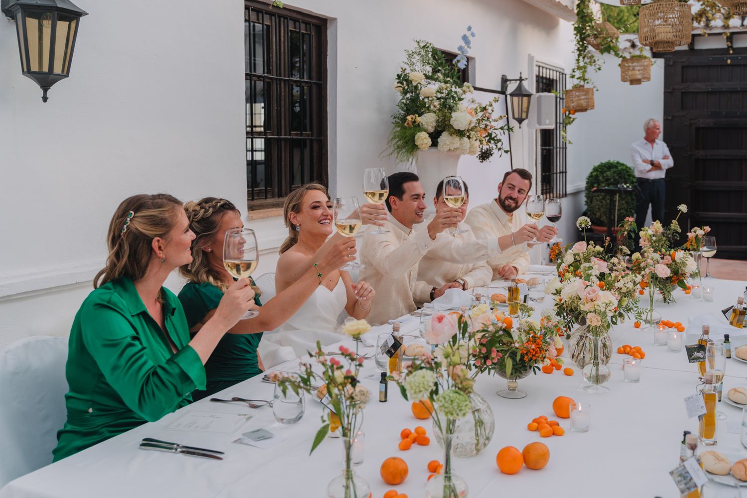 A festive outdoor dinner party with people toasting at a long white table decorated with citrus fruits and floral arrangements.