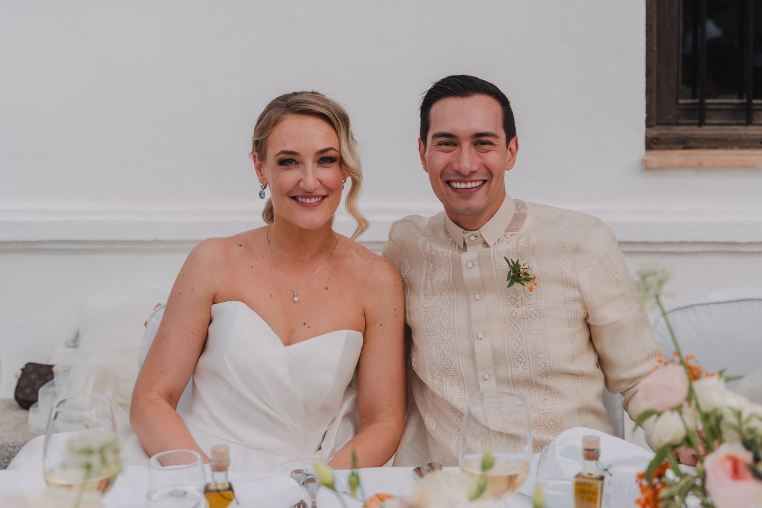 Wedding couple seated at reception table decorated with floral arrangements and white linens.