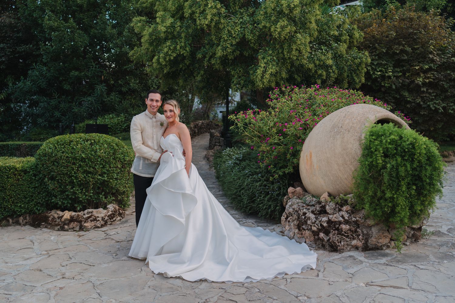 A couple poses in formal wedding attire next to a large decorative stone sphere with greenery in an outdoor garden.