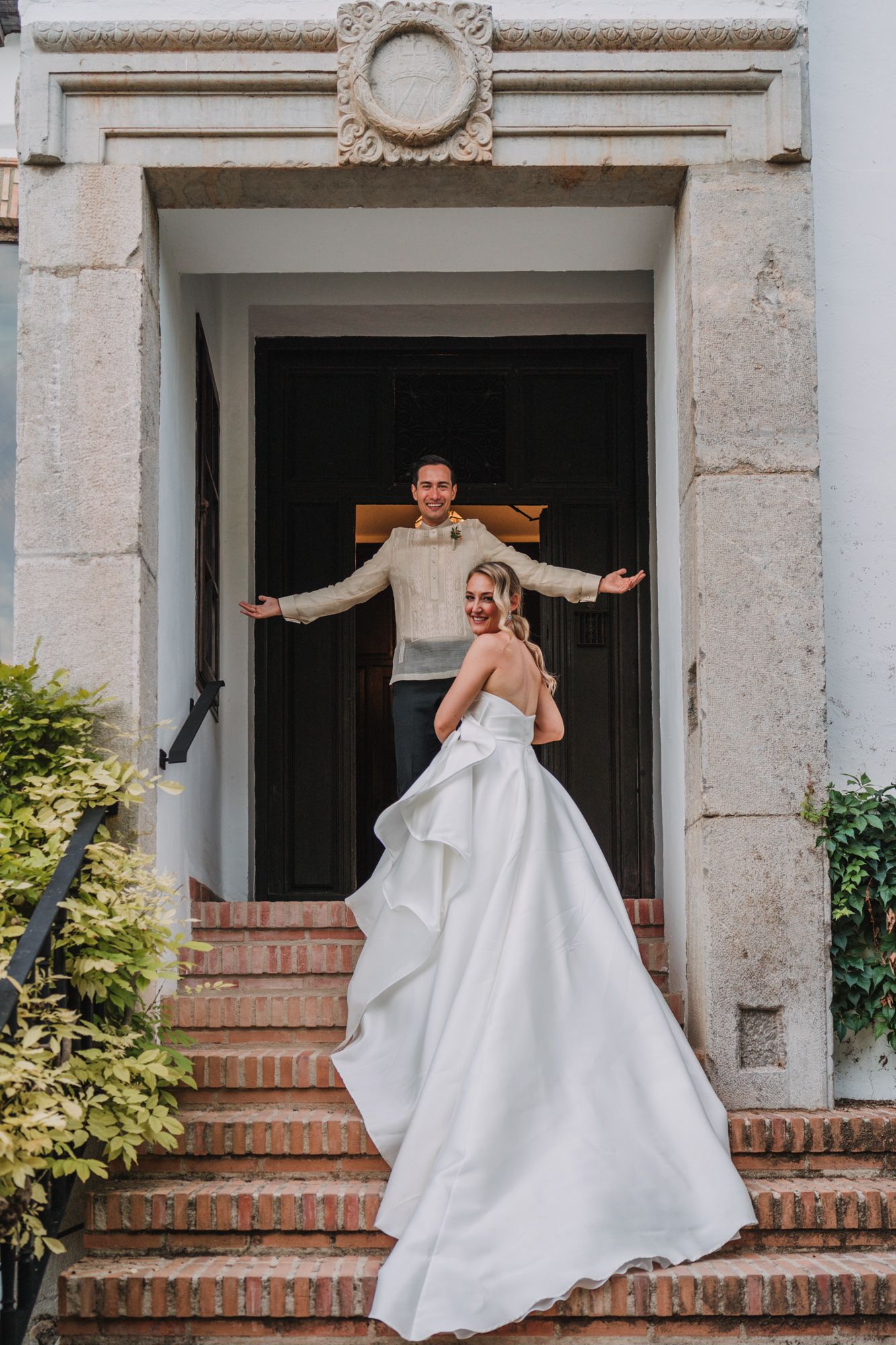 A wedding couple poses joyfully on elegant stone steps outside an ornate doorway of a historic building.