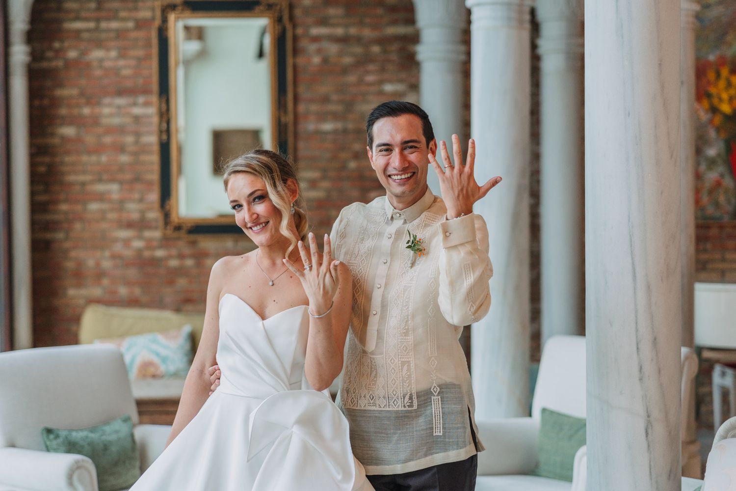 A joyful couple in wedding attire poses playfully indoors against a brick wall and white columns.