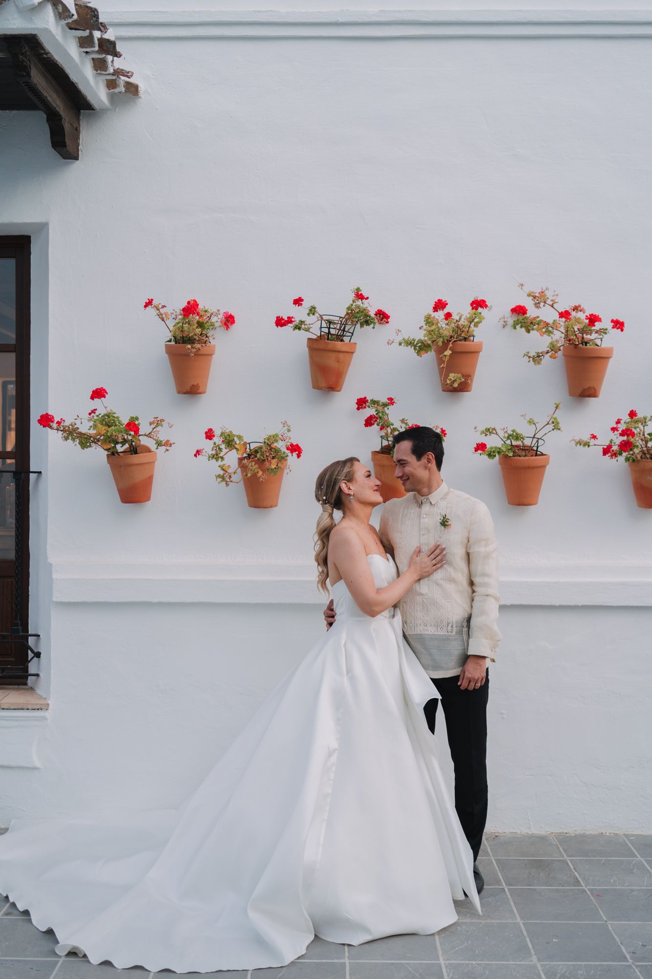 A romantic wedding moment against a white wall decorated with terracotta pots filled with red flowers.