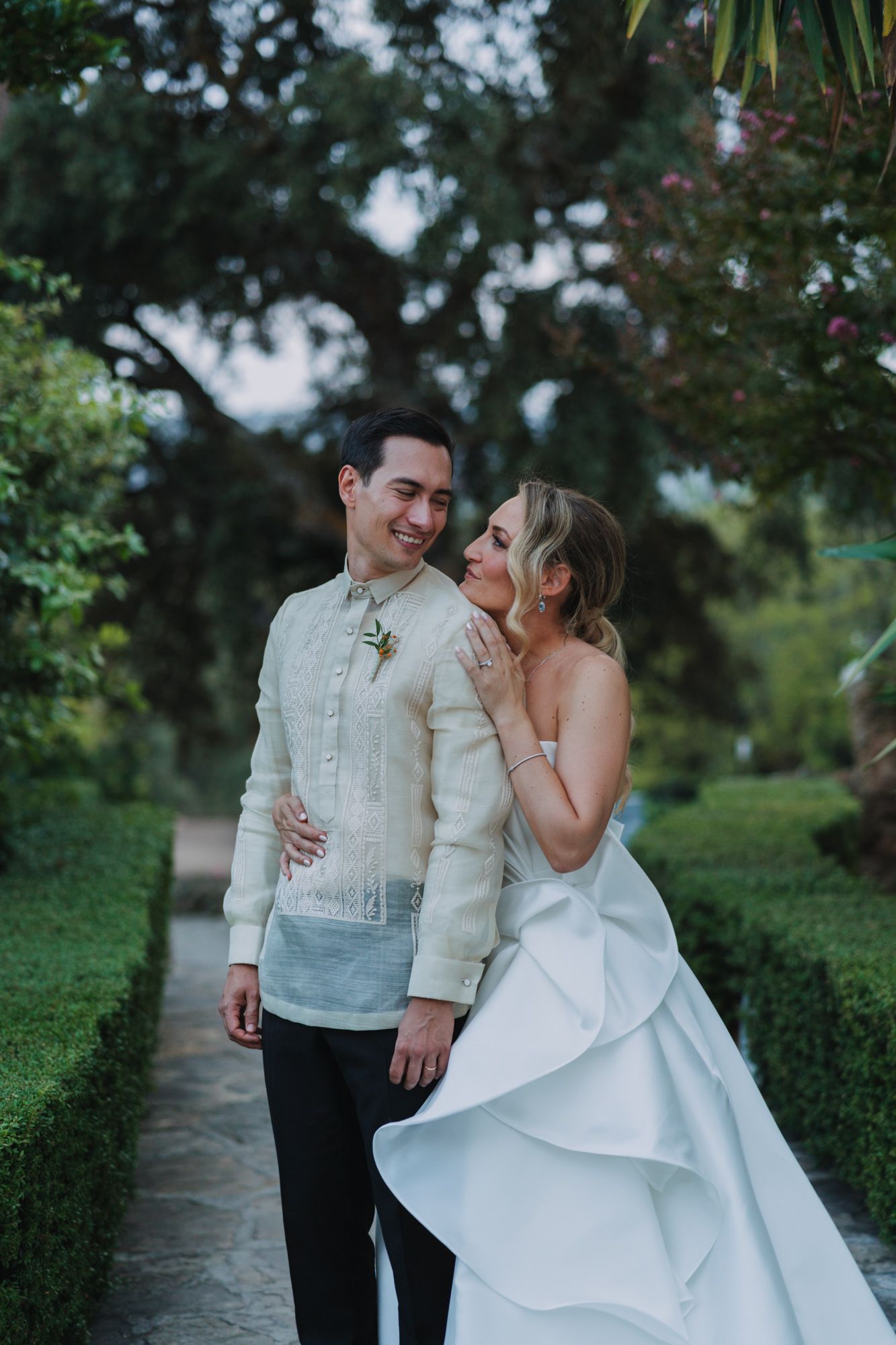 A romantic wedding portrait of a couple in traditional Filipino attire standing on a garden path with lush greenery.