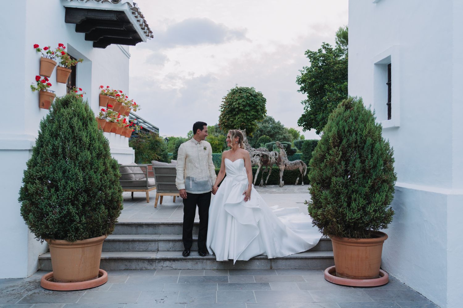 Couple standing between decorative potted topiaries on white building steps.