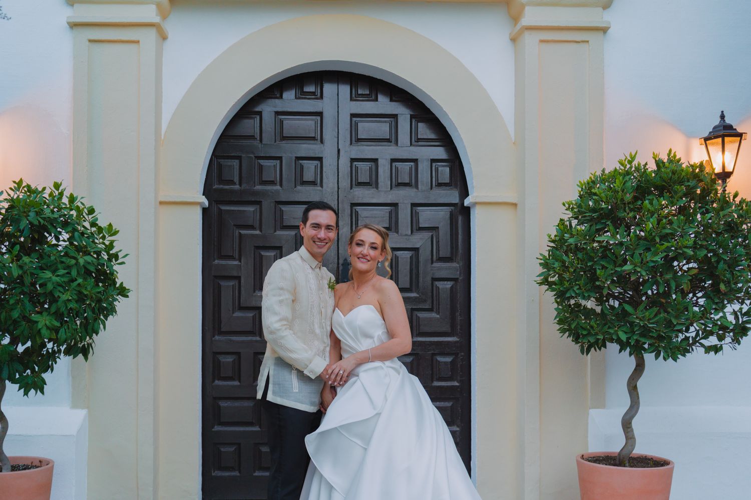 Wedding couple posing in archway with ornate dark wooden door and potted plants.