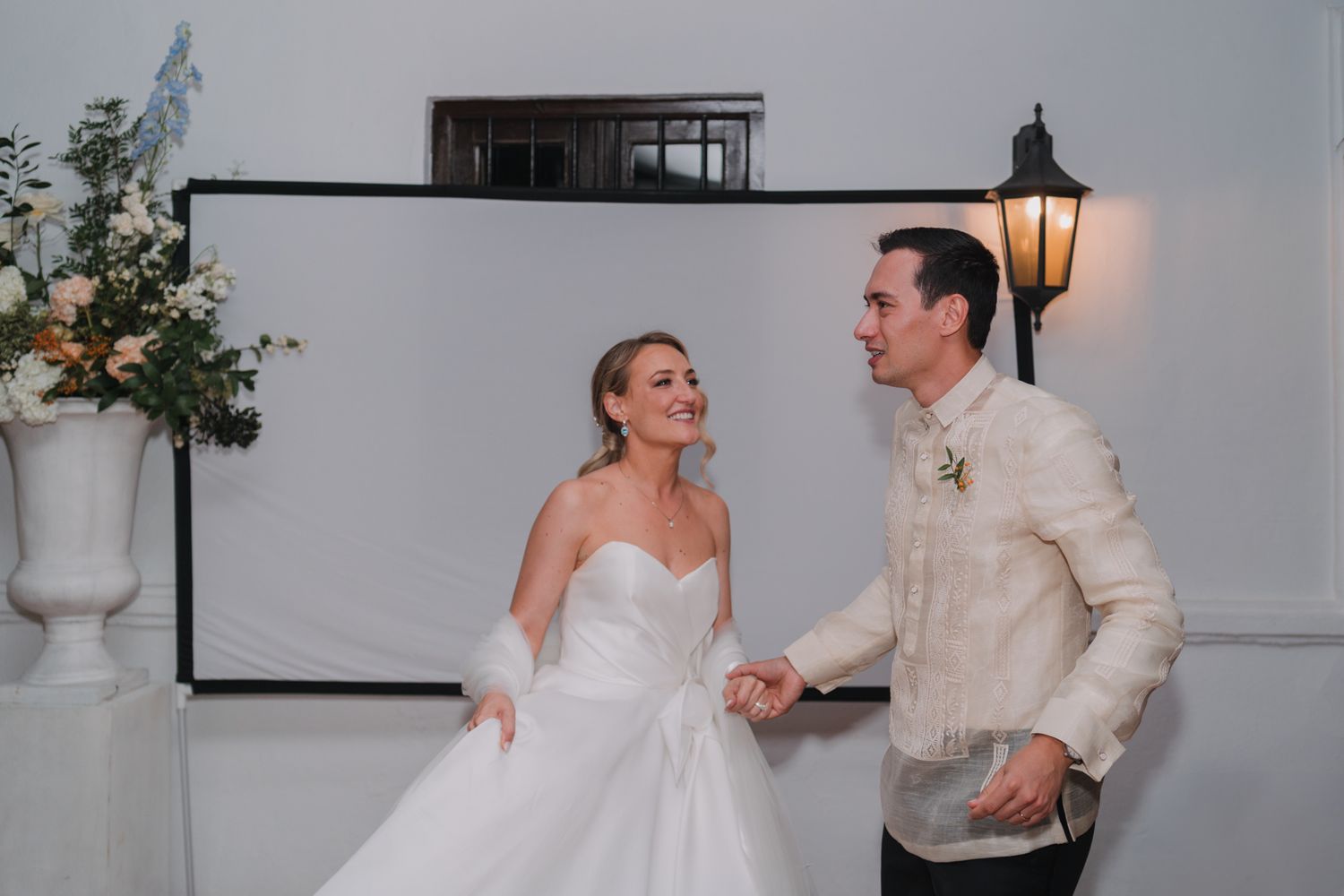 A joyful wedding couple shares a candid dancing moment in their elegant white attire next to a decorative floral arrangement.