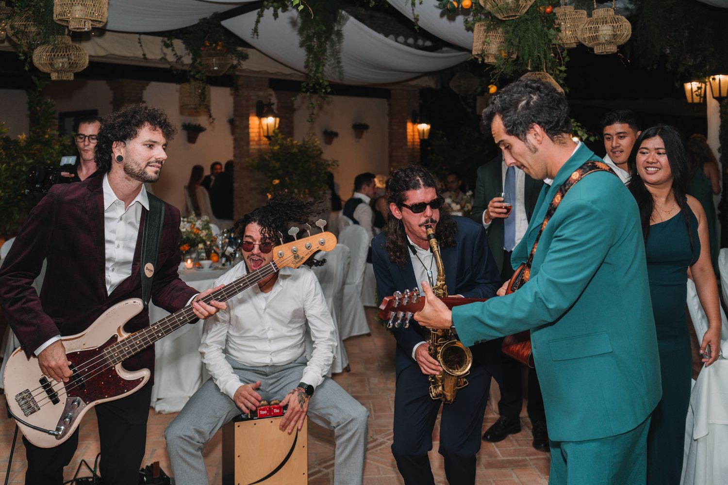Musicians perform energetically at a wedding reception with guitars and saxophone in an outdoor covered venue.