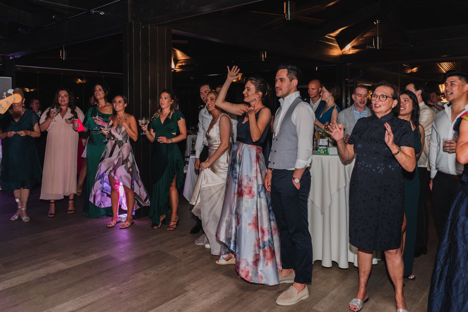 Wedding reception guests dance and celebrate together on a dimly lit dance floor.