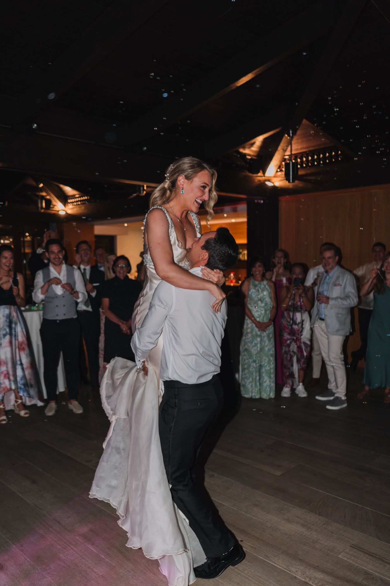 A wedding couple shares their first dance surrounded by guests in a dimly lit reception venue.
