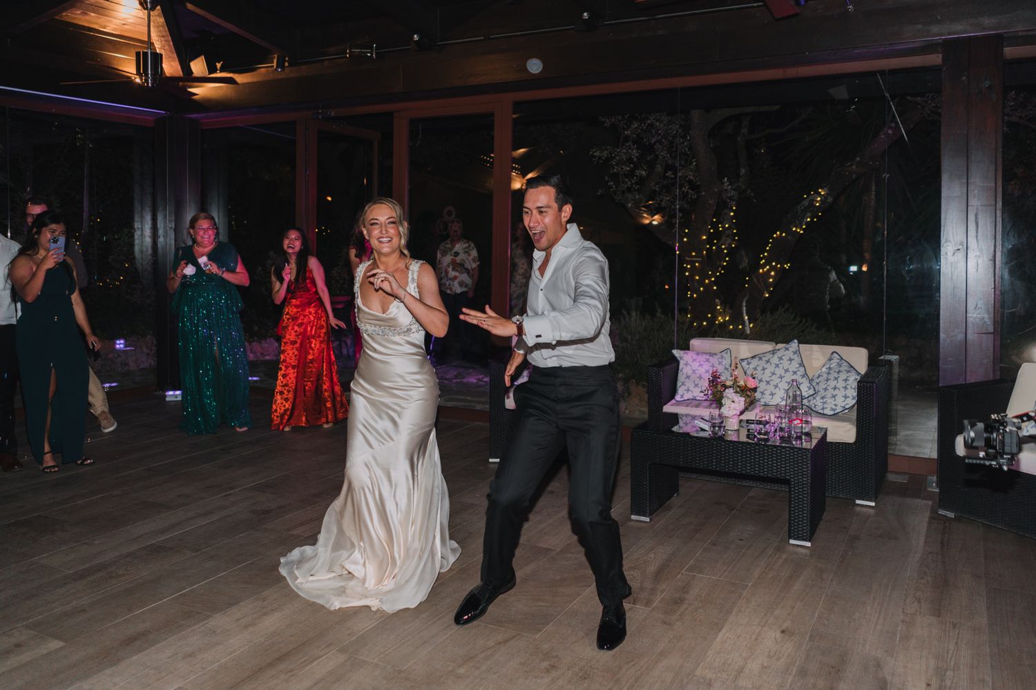 A newlywed couple dances energetically on a dark wooden floor at their wedding reception.