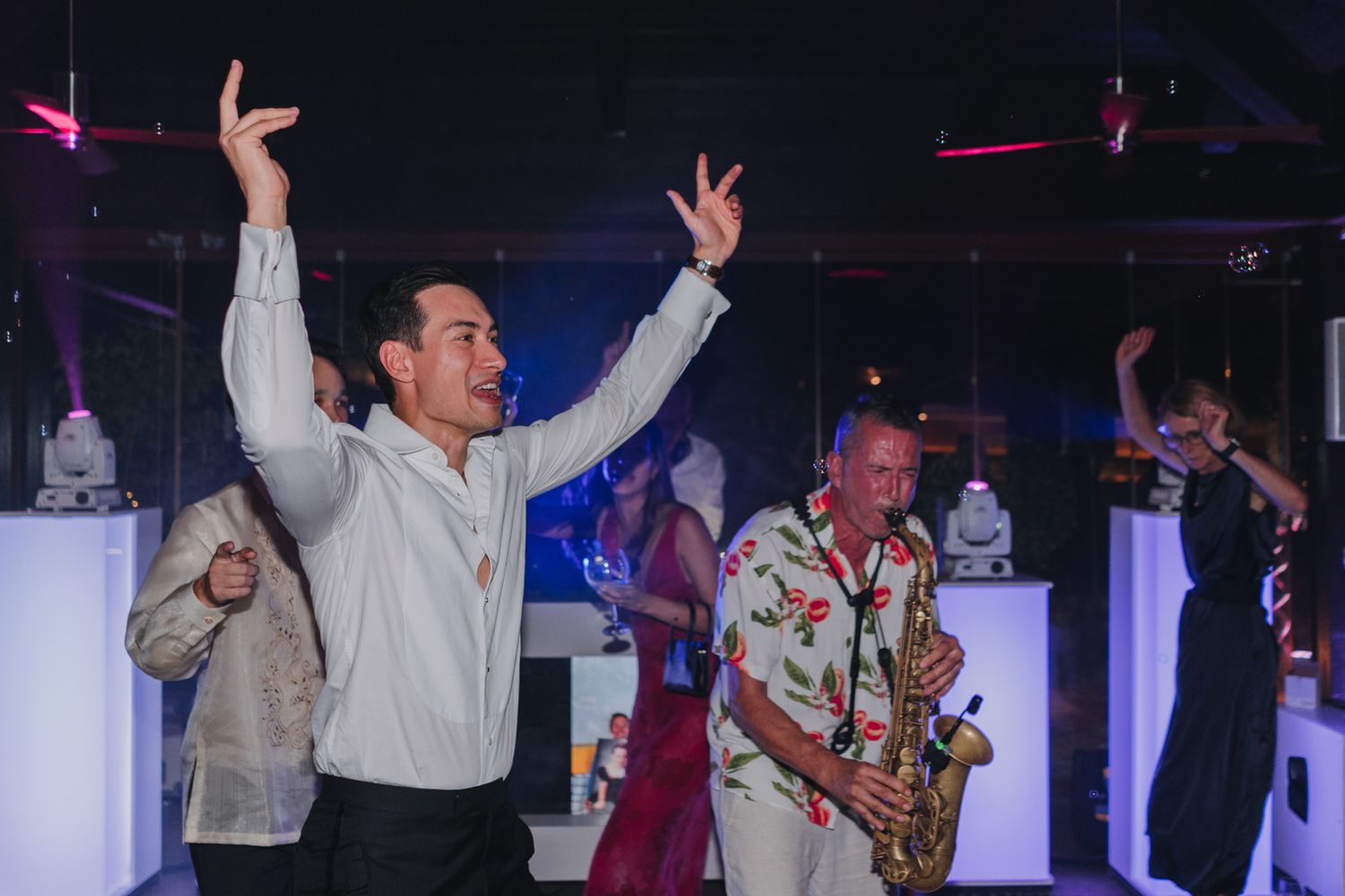 People dancing and celebrating at a nightclub with a saxophone player performing in colorful attire on stage.