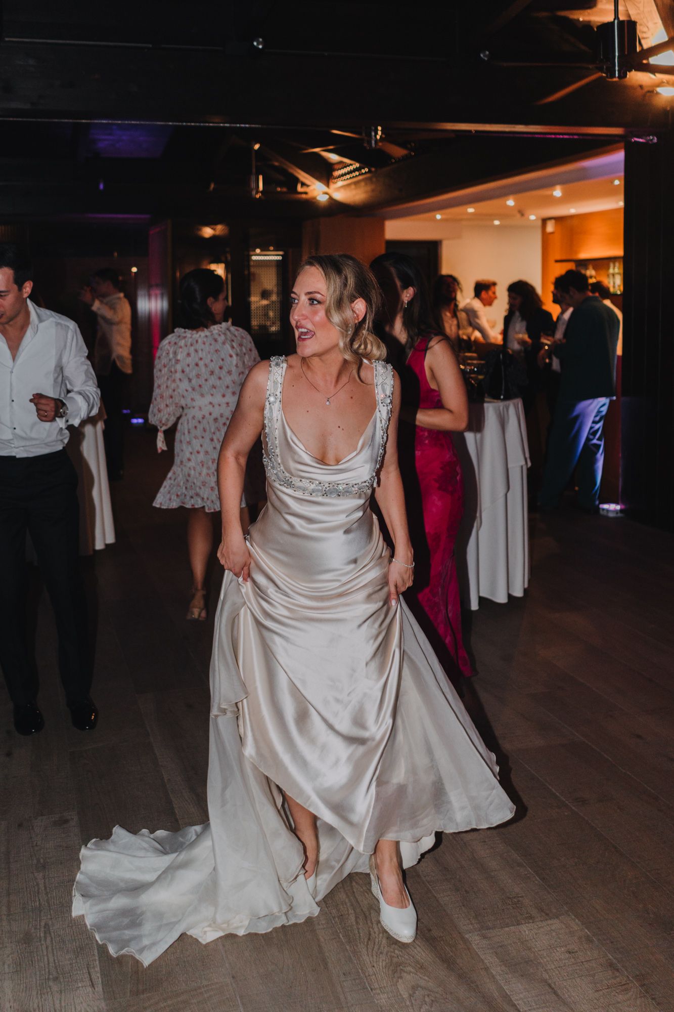 Person in flowing white satin wedding gown dances energetically on wooden floor at evening reception celebration.
