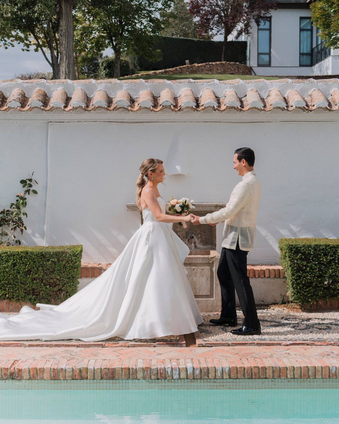 Two people in wedding attire stand near a pool beside a white stucco wall with Spanish clay roof tiles.