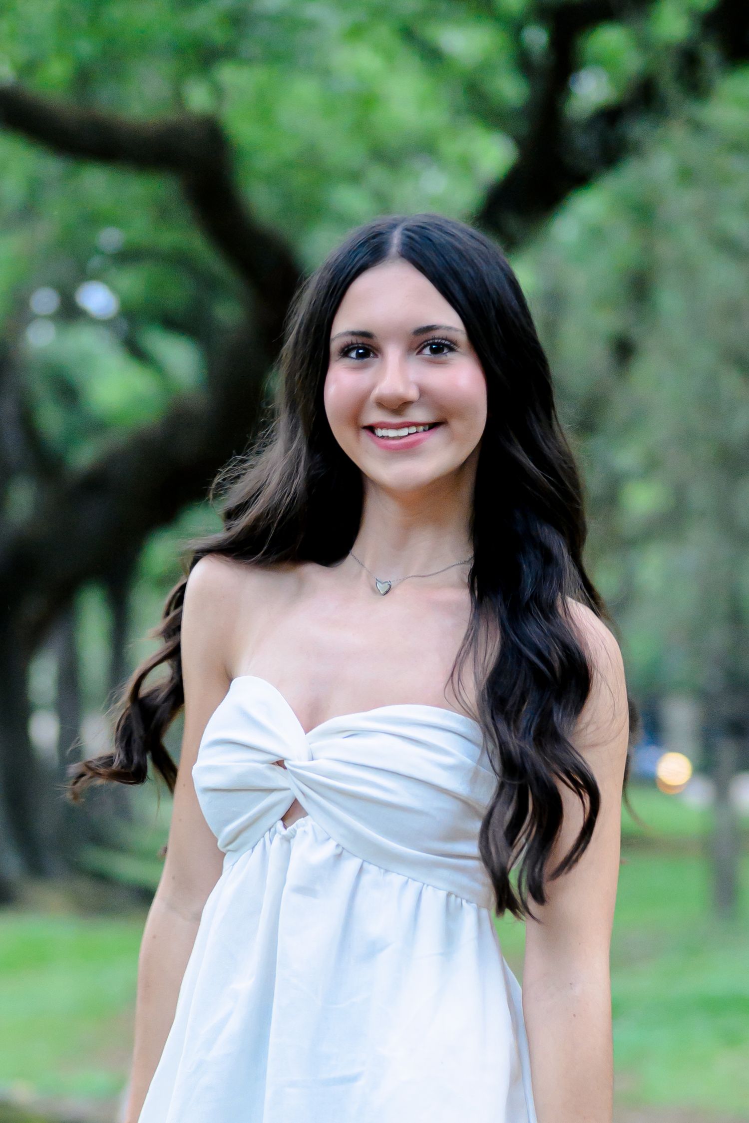 Portrait of someone in a strapless white summer dress standing outdoors with trees in a park setting.