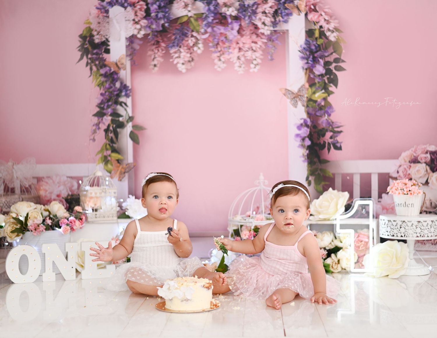 Pink floral cake smash setup with decorated arch, white lettering, and birthday props against a rosy backdrop.