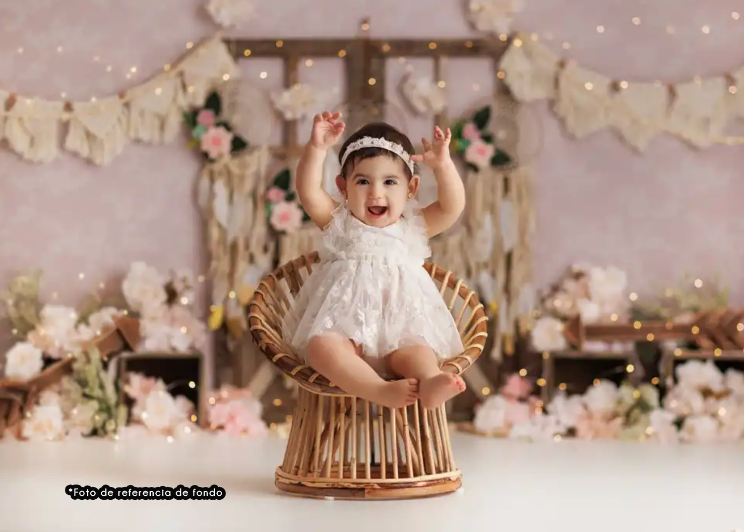 Baby in white dress sits on rattan chair surrounded by dreamy floral decorations and sparkly lights in pink studio setup.