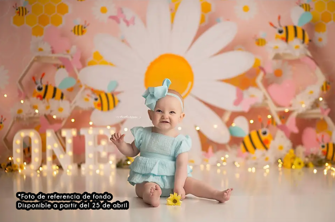 First birthday photo setup with daisy backdrop, illuminated ONE sign, and cute bee decorations in pink and yellow tones.