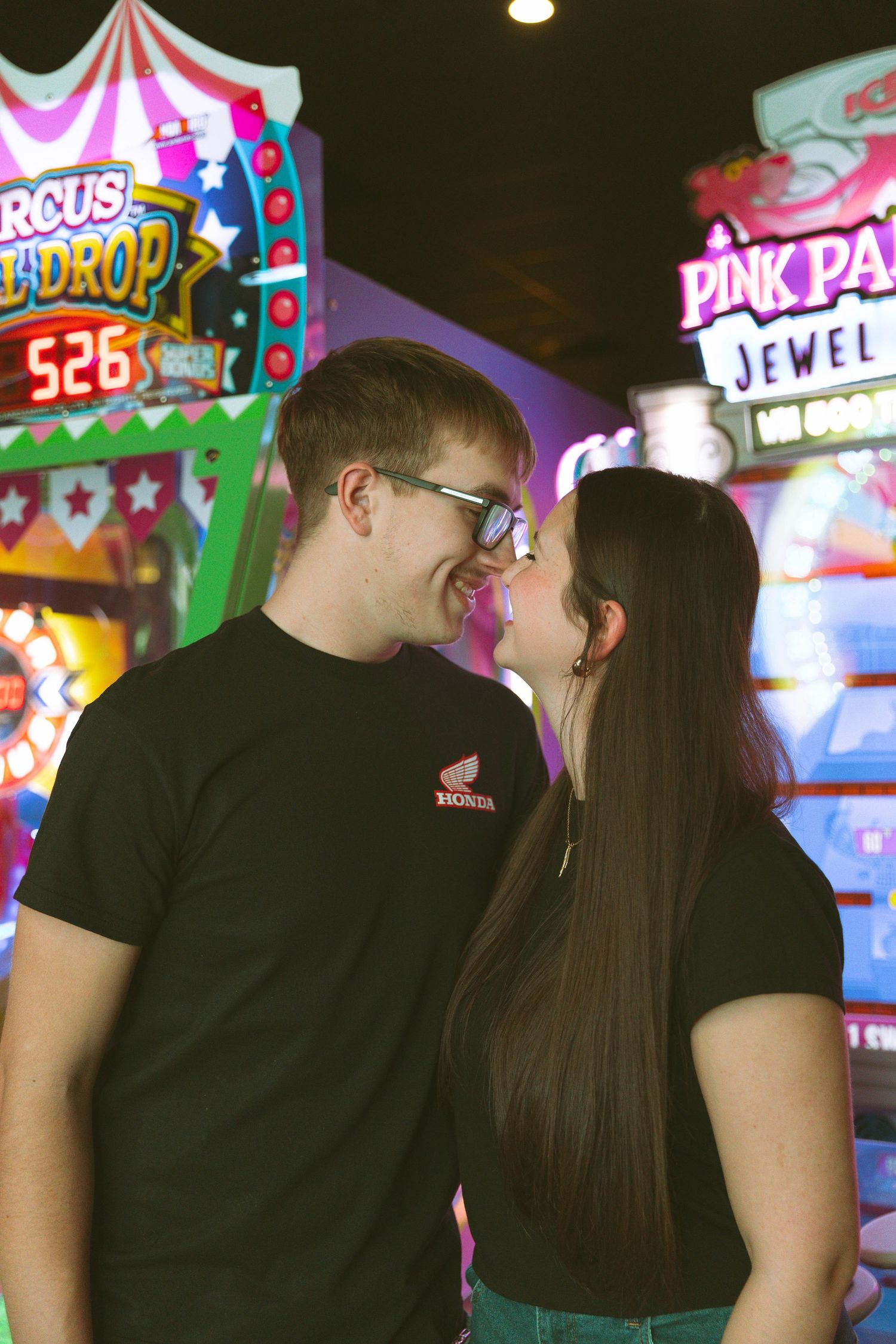 A couple shares a romantic moment in front of colorful arcade games with neon lights at an entertainment venue.
