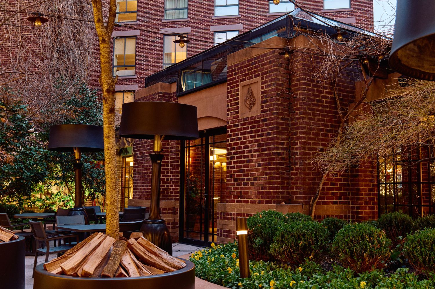 Cozy outdoor patio area with brick building and fire pit at dusk featuring warm lighting and landscaping.