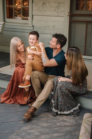 A family sits together on wooden steps wearing coordinated autumn-toned outfits in a casual outdoor setting.