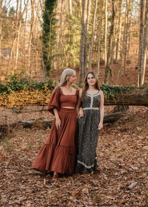 Two people in flowing dresses stand together in an autumn forest setting with fallen leaves.