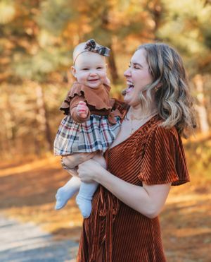 A baby in plaid dress and rust-colored top smiles while being held outdoors in autumn.