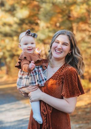 A baby in autumnal outfit smiles while being held outdoors against fall foliage.