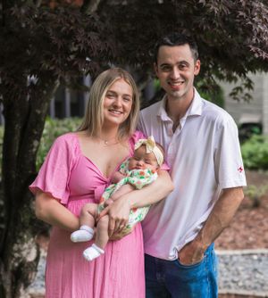 A couple stands outdoors with infant under trees in summer attire.