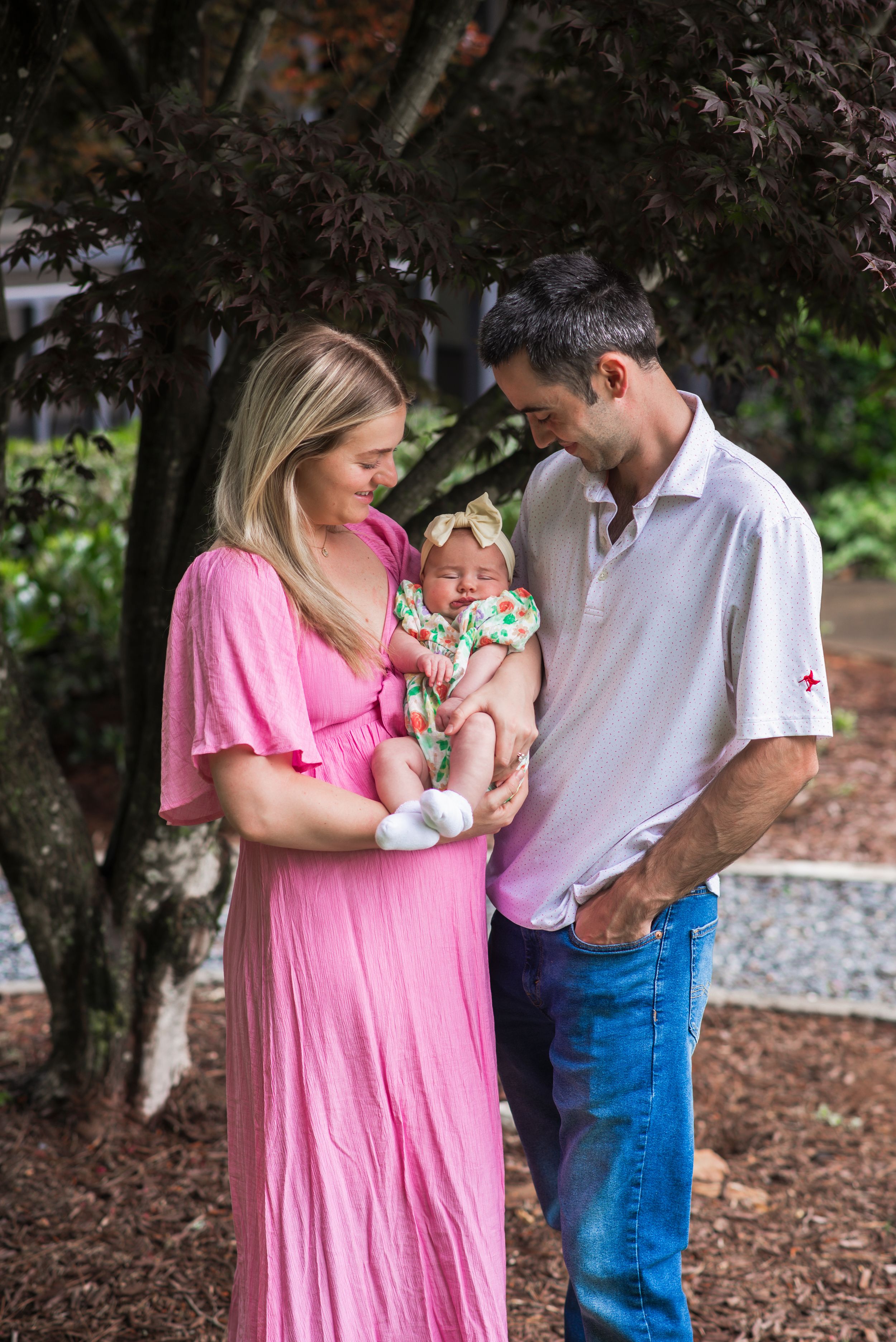 A couple in pink dress and white shirt share a tender moment with their baby under a tree.